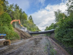 Two excavators tearing down a bridge on a gravel road, flanked by forest and a cloudy sky, bridge