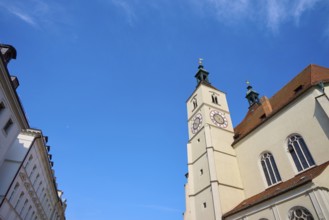 Gothic-style church tower and building under a bright blue sky, flanked by other buildings,