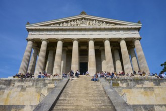 Temple-like structure with monumental columns and visitors on a staircase under a blue sky,