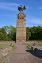 Monument with eagle on a pedestal, surrounded by trees. Wide paved path leads to the memorial.