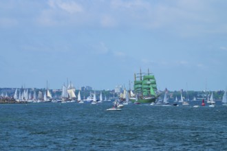 Green tall ship Alexander von Humbold 2 surrounded by many boats in front of an urban backdrop,