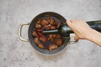 A hand pours red wine into the pot filled with seared chunks of beef and vegetables
