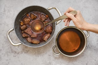 A person is using a ladle to add beef broth to a pot filled with fried chunks of beef shoulder and