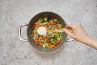 Freshly chopped carrots, celery, and onions are fried in a pot. A hand is sprinkling flour over the