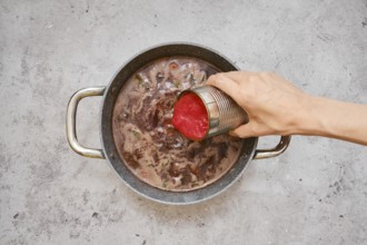 A hand pours diced tomatoes from a can into a pot filled with a rich, simmering beef stew