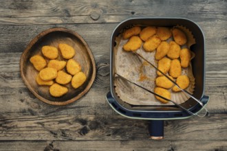 Golden and crispy chicken nuggets rest in an air fryer beside a wooden plate filled with freshly
