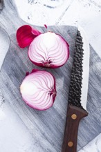 A vibrant red onion is cut in half, on a rustic wooden chopping board beside a sharp knife, ready