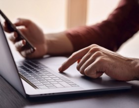 A man in a professional office environment multitasking with a smartphone and laptop, conveying