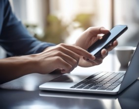A man in a professional office environment multitasking with a smartphone and laptop, conveying