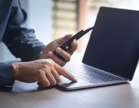 A man in a professional office environment multitasking with a smartphone and laptop, conveying