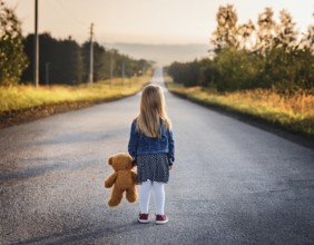 A young child stands alone holding a teddy bear in an urban outdoor setting, symbolizing innocence