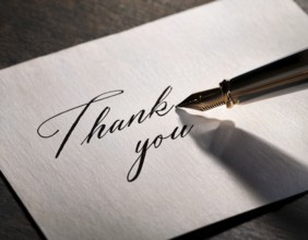 Elegant pen rests alongside a handwritten Thank You card on a wooden surface in soft light.
