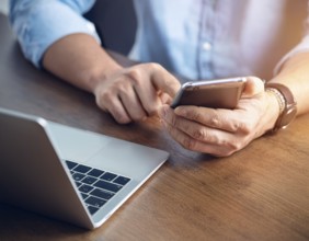 A man in a professional office environment multitasking with a smartphone and laptop, conveying