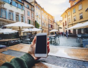 A person holding a smartphone at an outdoor cafe amidst a vibrant and lively urban setting. The