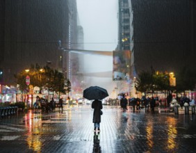 A solitary figure with an umbrella stands amidst a bustling city illuminated by lights, with