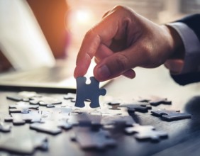 A person's hand placing a puzzle piece on a reflective surface in an office setting as the sun