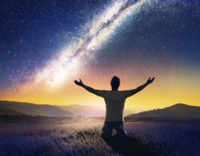 A man kneels in peaceful reflection under the vibrant Milky Way galaxy at sunset in an open natural