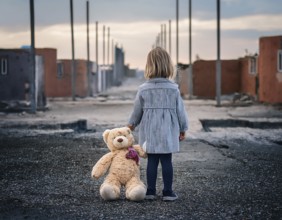 A young child stands alone holding a teddy bear in an urban outdoor setting, symbolizing innocence