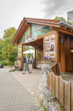 A wooden restaurant at the edge of the forest with passing hikers on a paved path, Lichtenstein