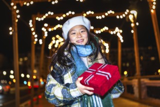 Smiling young woman holding christmas present enjoying christmas lights decoration at night