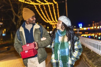 Young multiethnic couple walking arm in arm at illuminated christmas market, man carrying christmas
