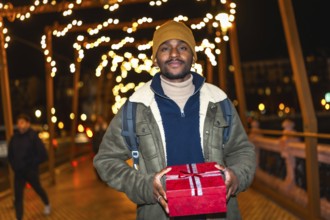 Young man holding christmas gift while walking on a city bridge decorated with christmas lights