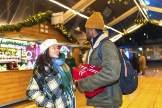 Young multiethnic couple exchanging christmas gifts at a christmas market, enjoying the festive