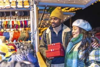 Young multiethnic couple enjoying christmas shopping at a decorated christmas market, holding red