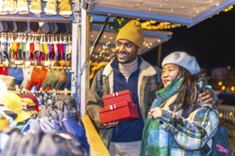 Smiling multiethnic couple holding christmas gifts while enjoying christmas market at night