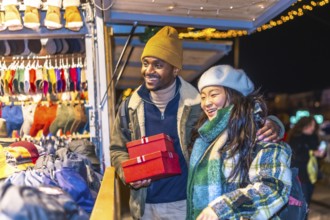 Multiethnic couple enjoying christmas shopping at a decorated christmas market, holding red gift
