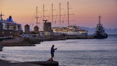 A fisherman fishing at sunset at an illuminated harbour with several ships on the horizon, Rhodes