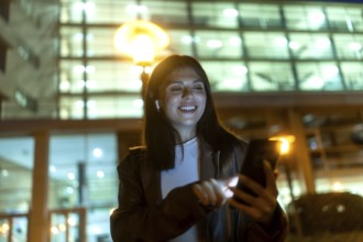 Young woman smiling and engaging with her smartphone, wearing wireless earbuds for connectivity and