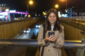 Young woman smiling while looking at her smartphone, standing on an urban bridge with city lights