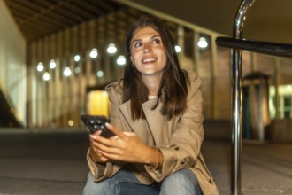Young woman smiling while holding a mobile phone, looking up and away, sitting in an urban
