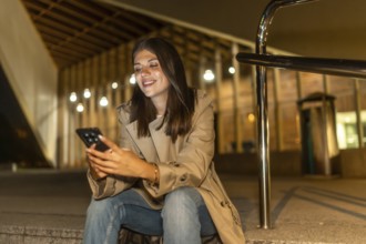 Young woman in a trench coat smiling, sitting on steps in an urban environment at night, connecting