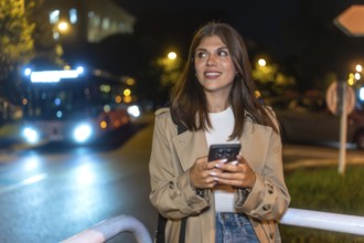 Young woman smiling while checking her mobile phone at a city bus stop at night, anticipating
