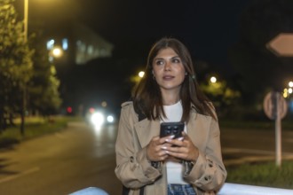 Young woman standing on city street at night, holding smartphone and looking away as warm car