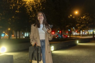 Woman in trench coat walking through an illuminated city park at night, smiling and confident while