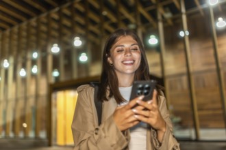 Young woman in a trench coat smiling, checking her smartphone, and experiencing happy moments while