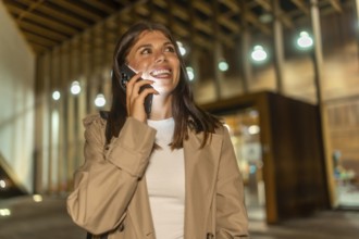 Young woman in trench coat smiling during a phone call, smartphone glow lighting her face on a