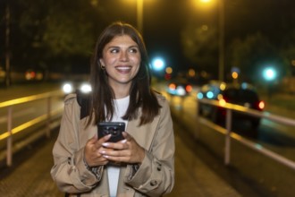 Young woman smiling while holding a smartphone and walking through a downtown street at night,