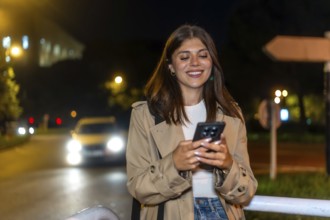Young brunette woman standing on a city street at night, smiling and checking her smartphone in a