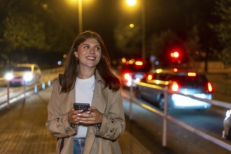 Young woman in a trench coat smiling, holding a phone, and looking away while walking on a city