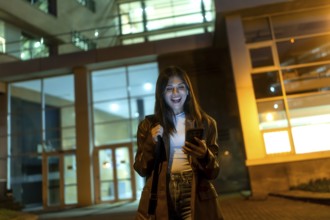 Young woman standing on an urban street at night, happily looking at her smartphone while holding a
