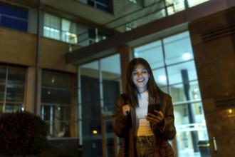 Young woman standing on a city street at night, looking at her smartphone and smiling with eyes