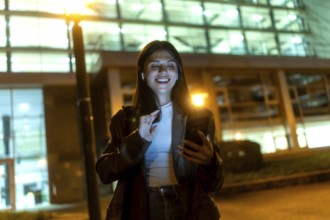 Young woman smiling broadly, wearing wireless earbuds and dark jacket, interacting with a