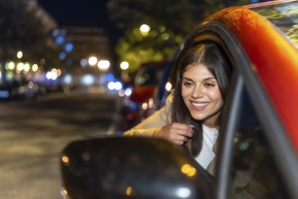 Young woman leaning out of a car window at night, smiling as city lights and bokeh blur by,