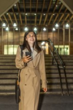Young woman dressed in a trench coat smiling, holding a mobile phone, and walking up illuminated
