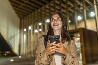 Young woman smiling, holding phone while walking illuminated city streets at night, confidently