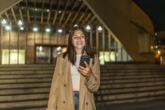 Young woman smiling while looking at her mobile phone outdoors, standing at night in front of a